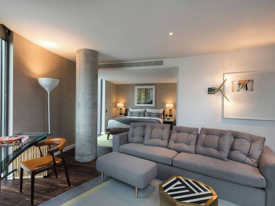 Una habitación de hotel moderna con un sofá gris, una mesa de vidrio y una silla. Hay una cama con cabecera y una mesita de noche con una lámpara. La habitación tiene un ventanal grande y vistas de la ciudad.
