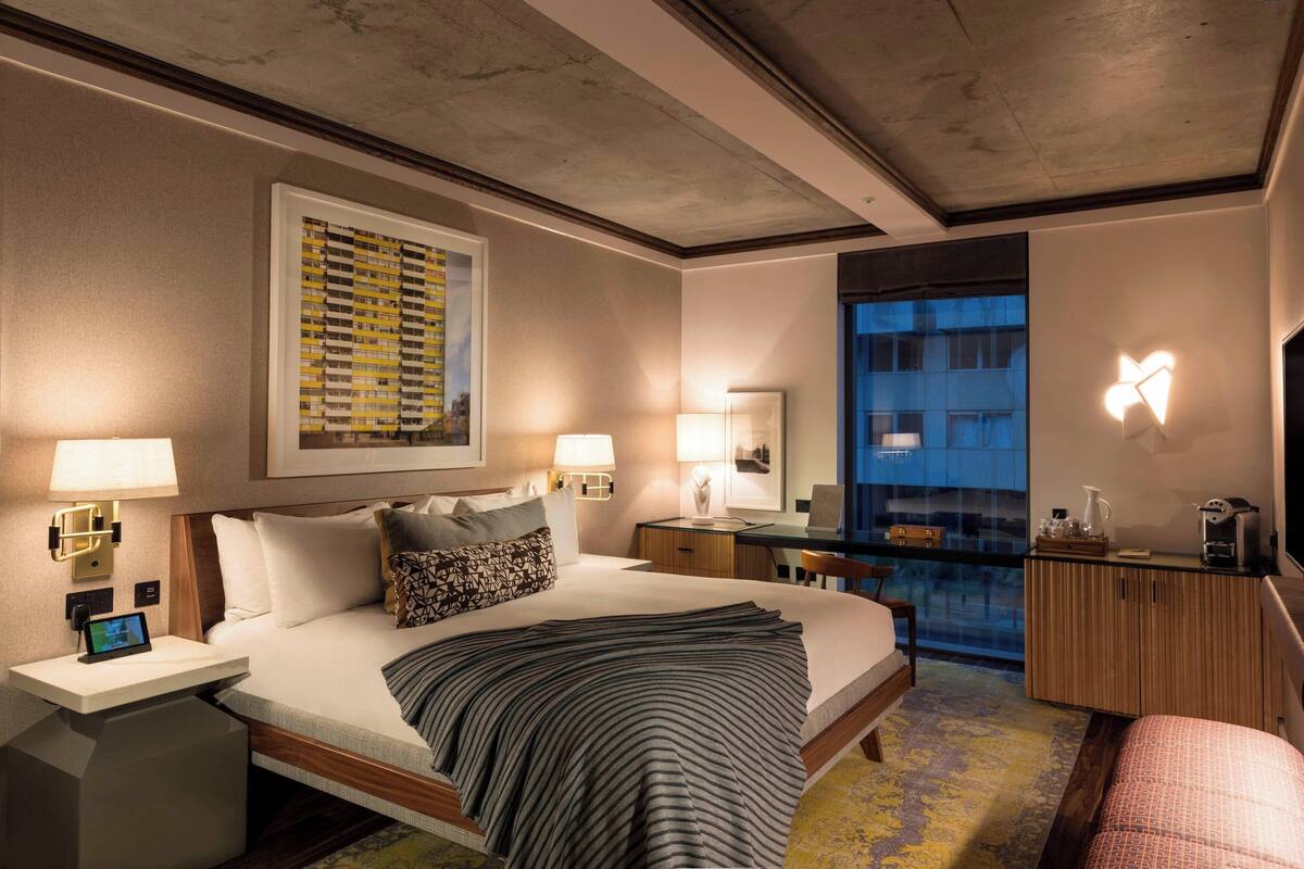 A hotel room with a large bed, bedside tables, a desk, a chair, a mirror, and a view of a building through the window.