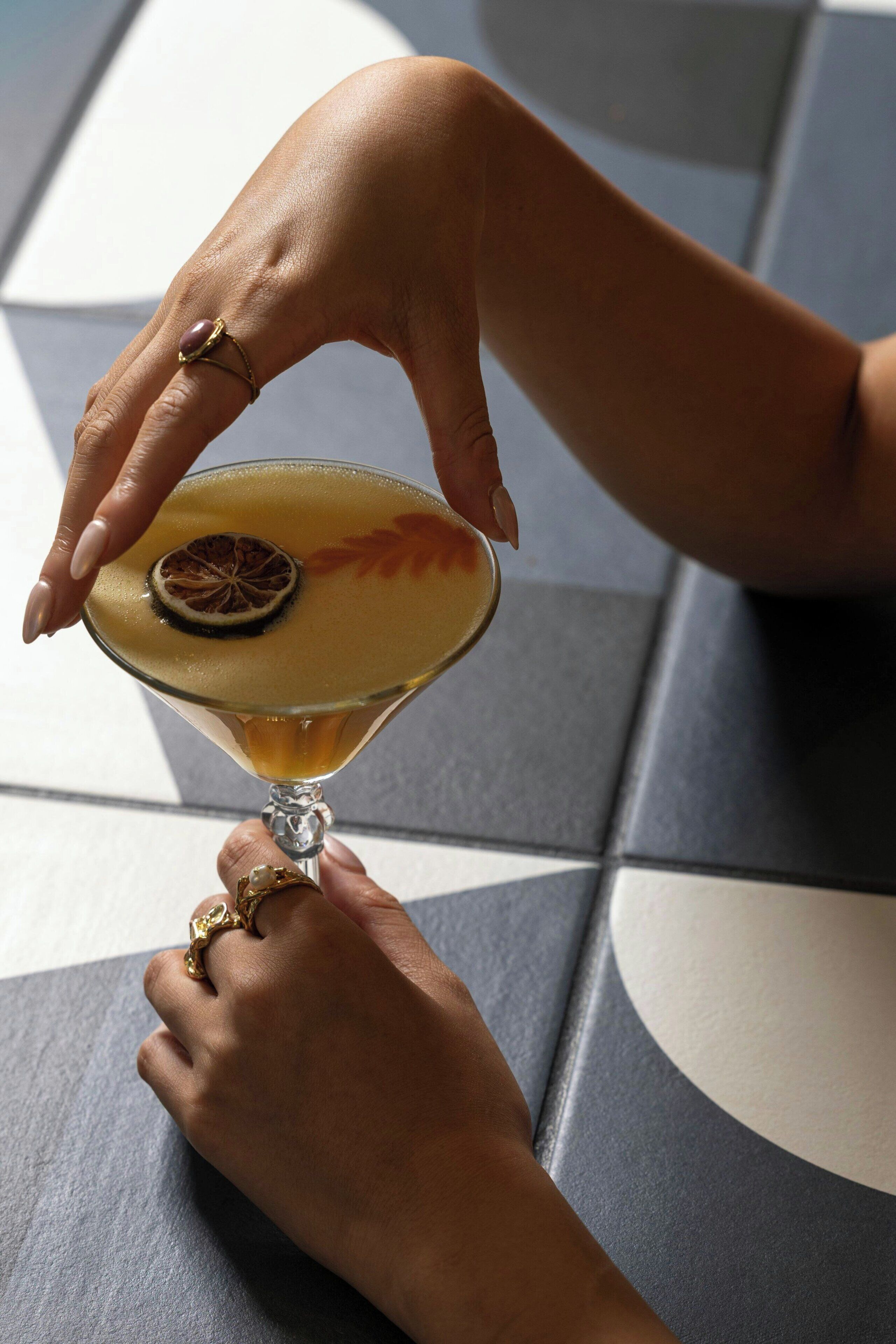 Une personne tenant un verre à martini, agrémenté d’une garniture, assise à une table posée sur un sol carrelé.