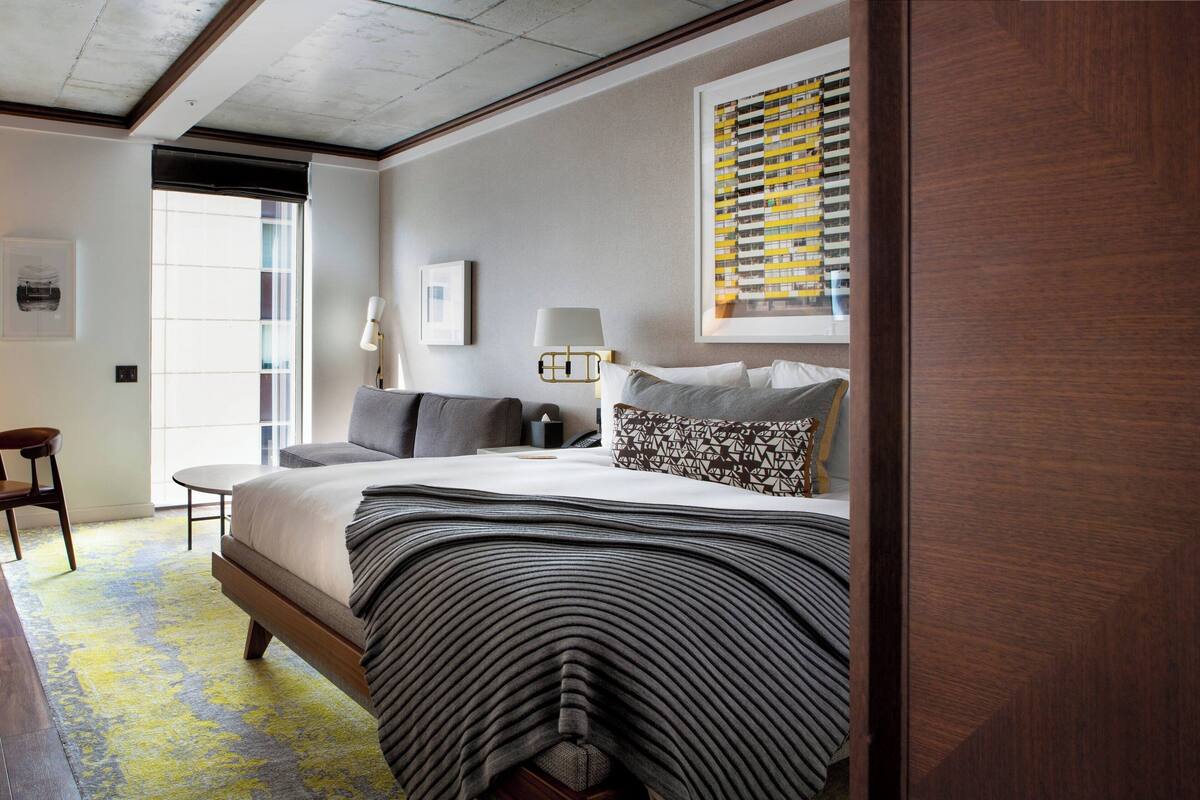 A modern hotel room with a large bed, a bedside table, a lamp, and a window with blinds.