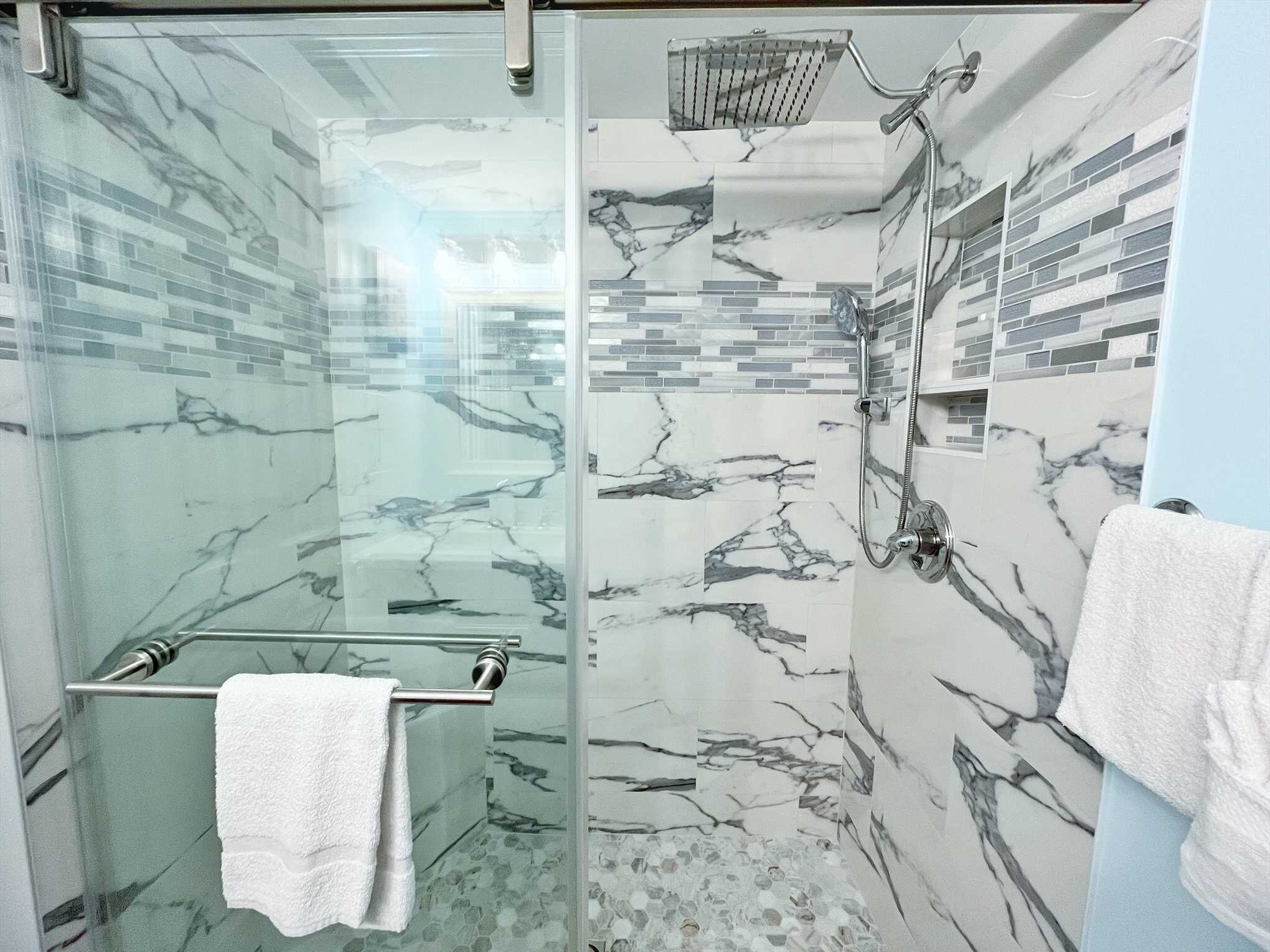 Ein modernes Badezimmer mit Duschkabine aus Glas, einem Waschtisch ebenfalls mit Glasabdeckung und einem Handtuch, sowie Wänden mit Marmor-Optik.