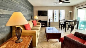 A living room with a sofa, coffee table, and TV. There is a dining area with a table and chairs. The room has a ceiling fan and a window with blinds.