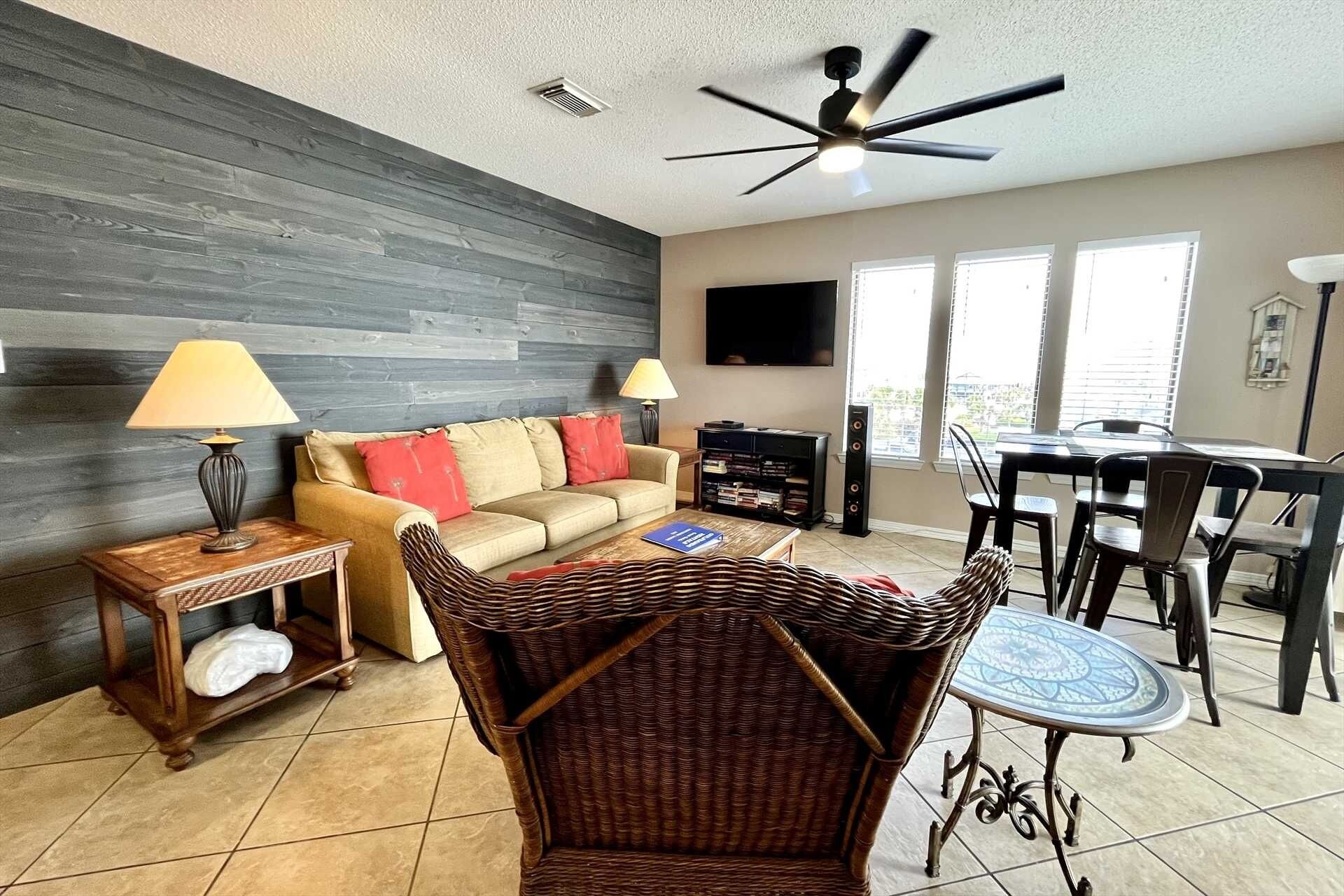 A living room with a sofa, two lamps, a television, a dining area with chairs and a table, and a ceiling fan.