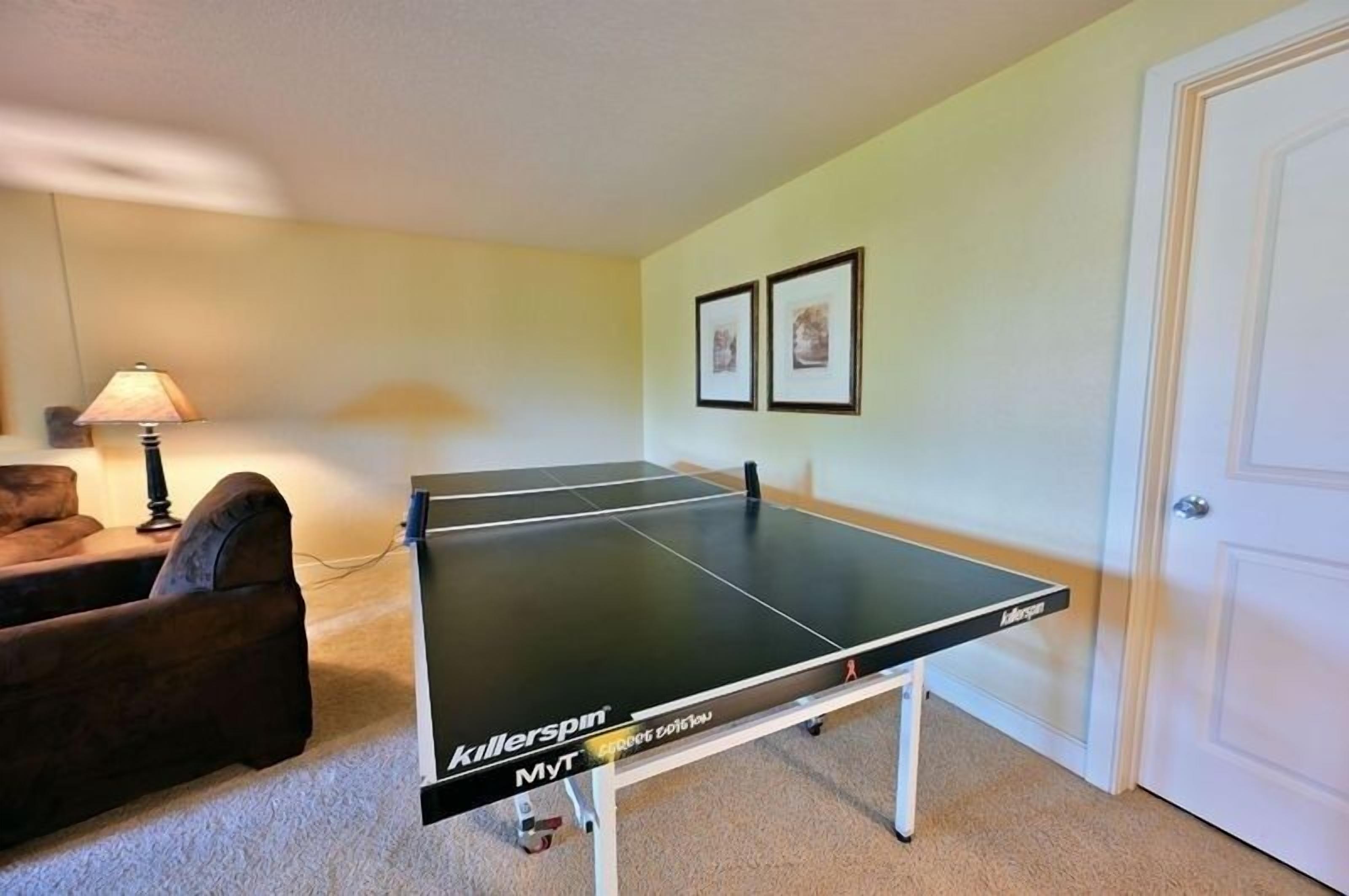 A ping pong table in a room with a carpeted floor, a lamp, and two framed pictures on the wall.