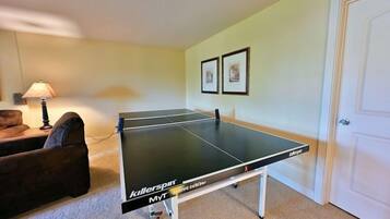 A ping pong table in a room with a carpeted floor, a lamp, and two framed pictures on the wall.