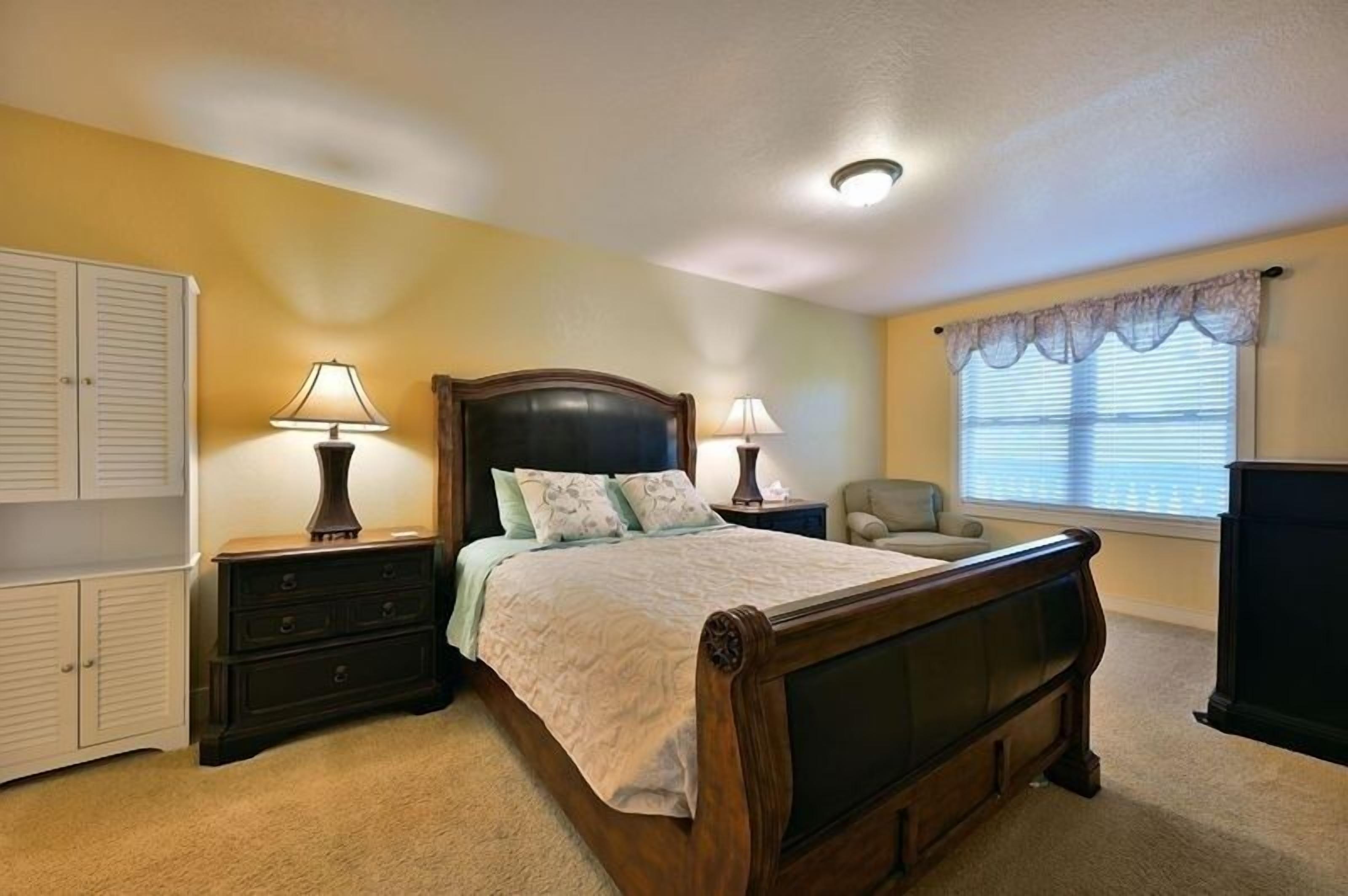 Une chambre à coucher comprenant un lit, des tables de chevet, une chaise, une armoire et une fenêtre avec des stores.
