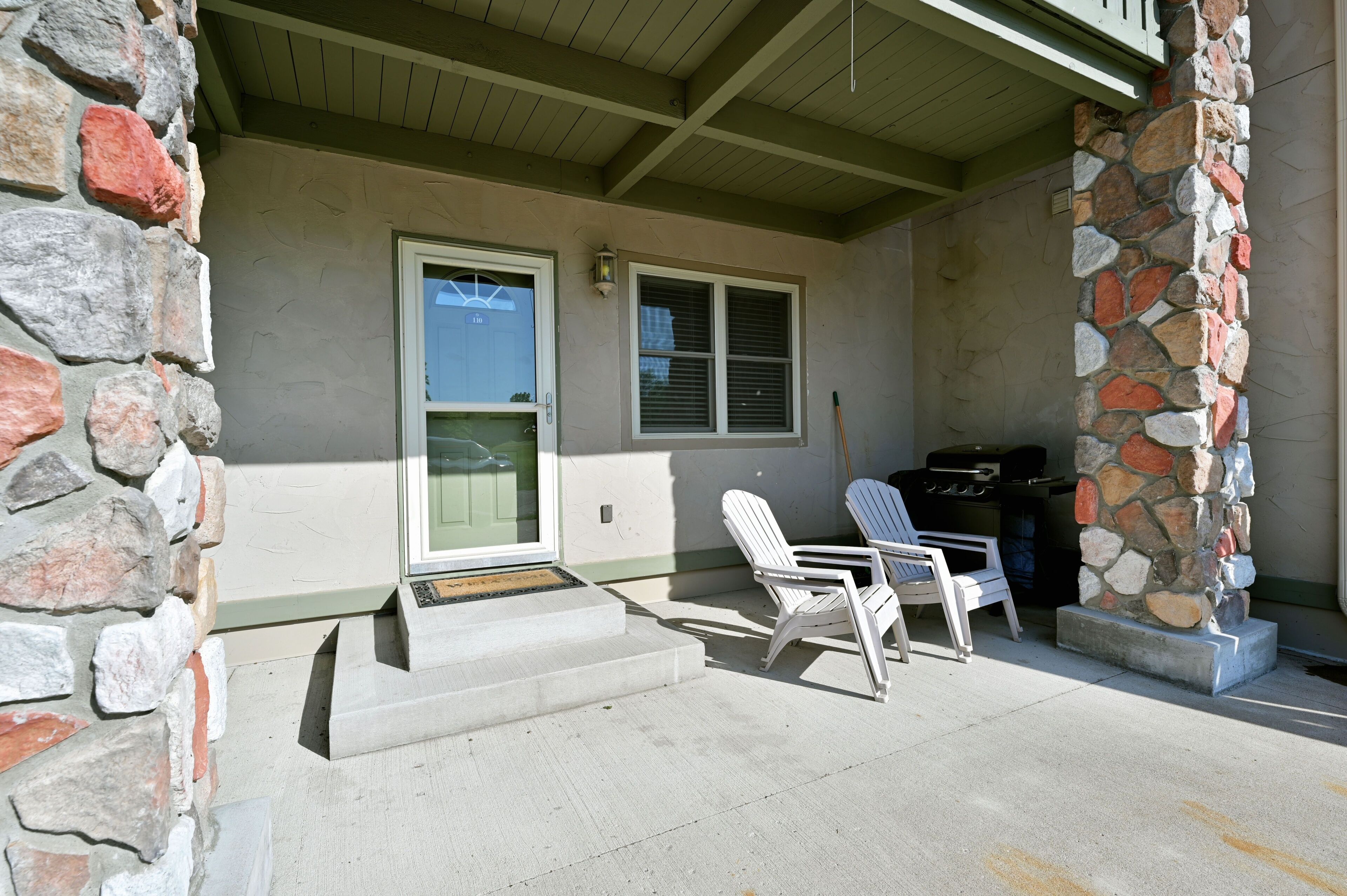 A covered porch with stone walls, a sliding glass door, and two white Adirondack chairs.