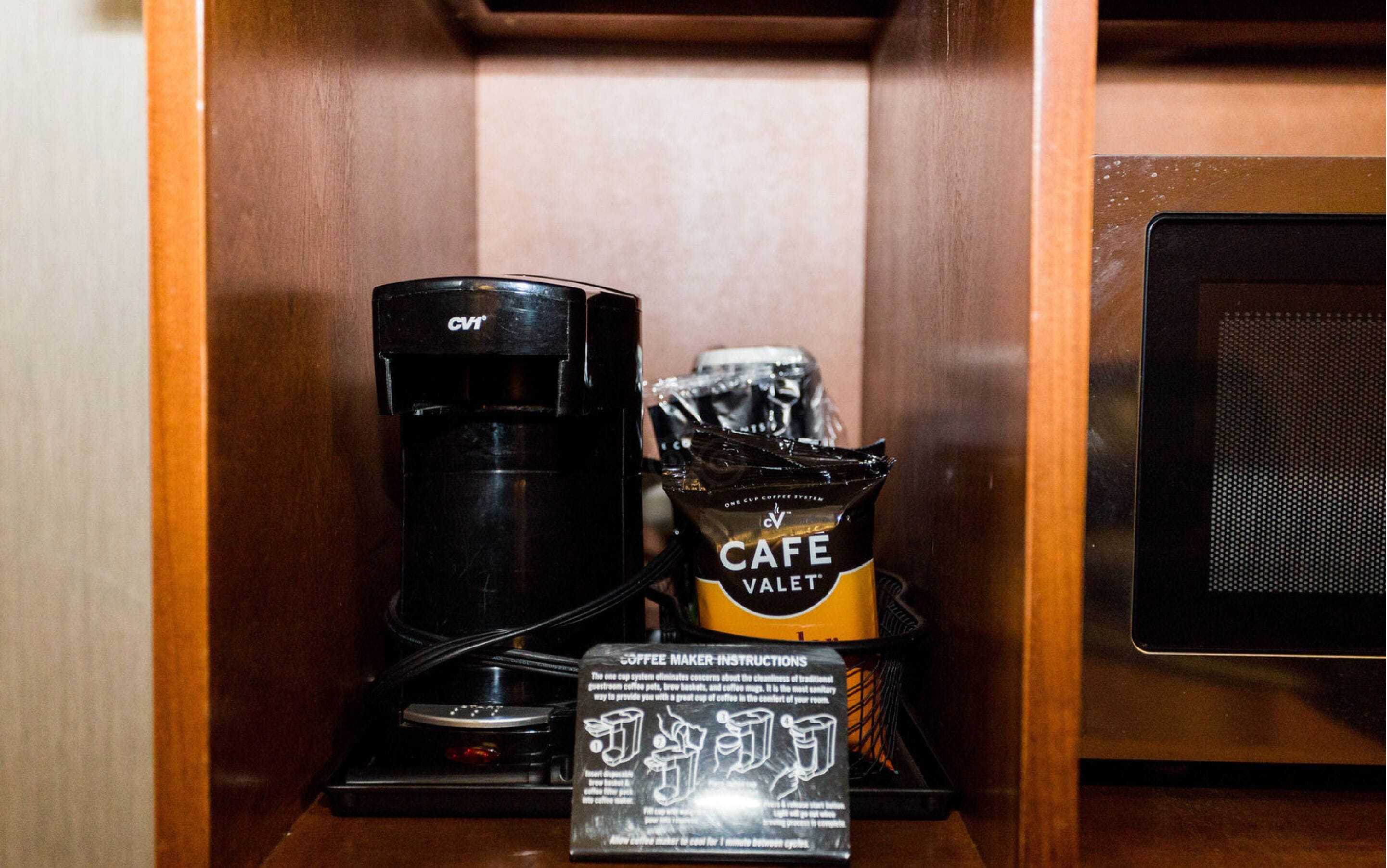 A coffee maker, a bag of Cafe Valet coffee pods, and a coffee maker instruction manual in a wooden cabinet.