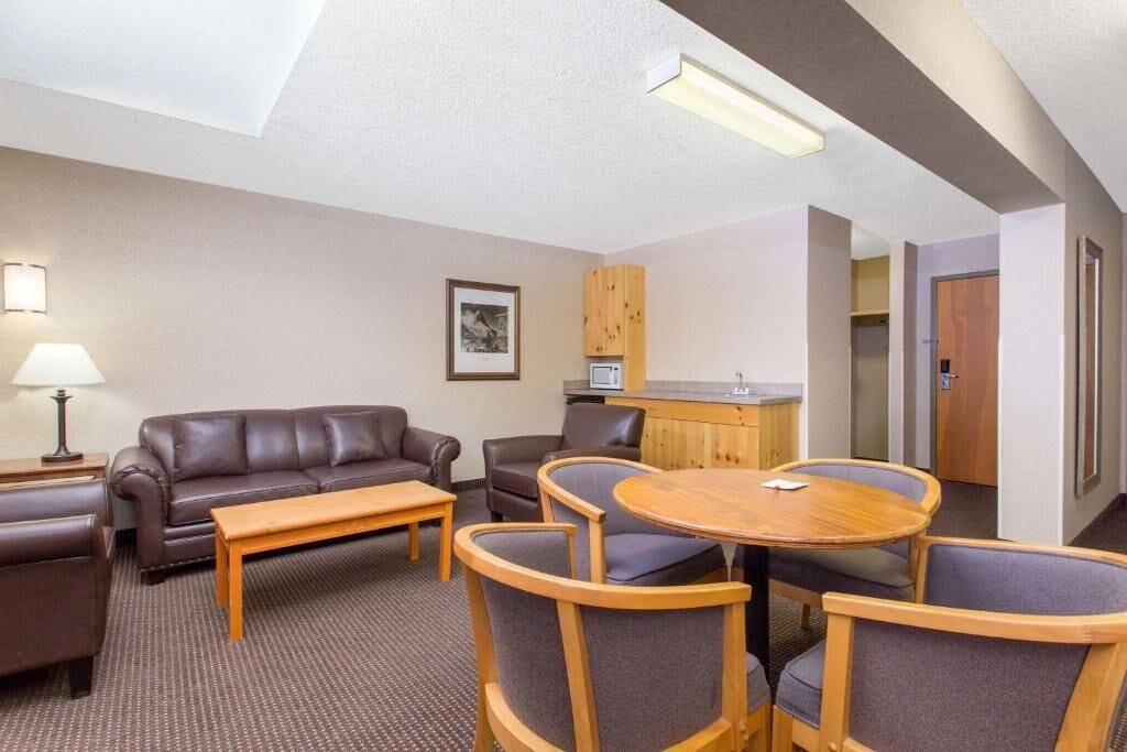 A hotel room with a sofa, two armchairs, a wooden coffee table, a lamp, a framed picture on the wall, and a kitchenette area.