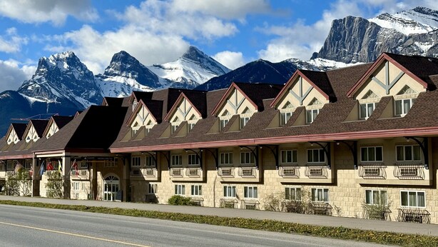 A row of chalet-style buildings with balconies, set against a backdrop of snow-capped mountains.