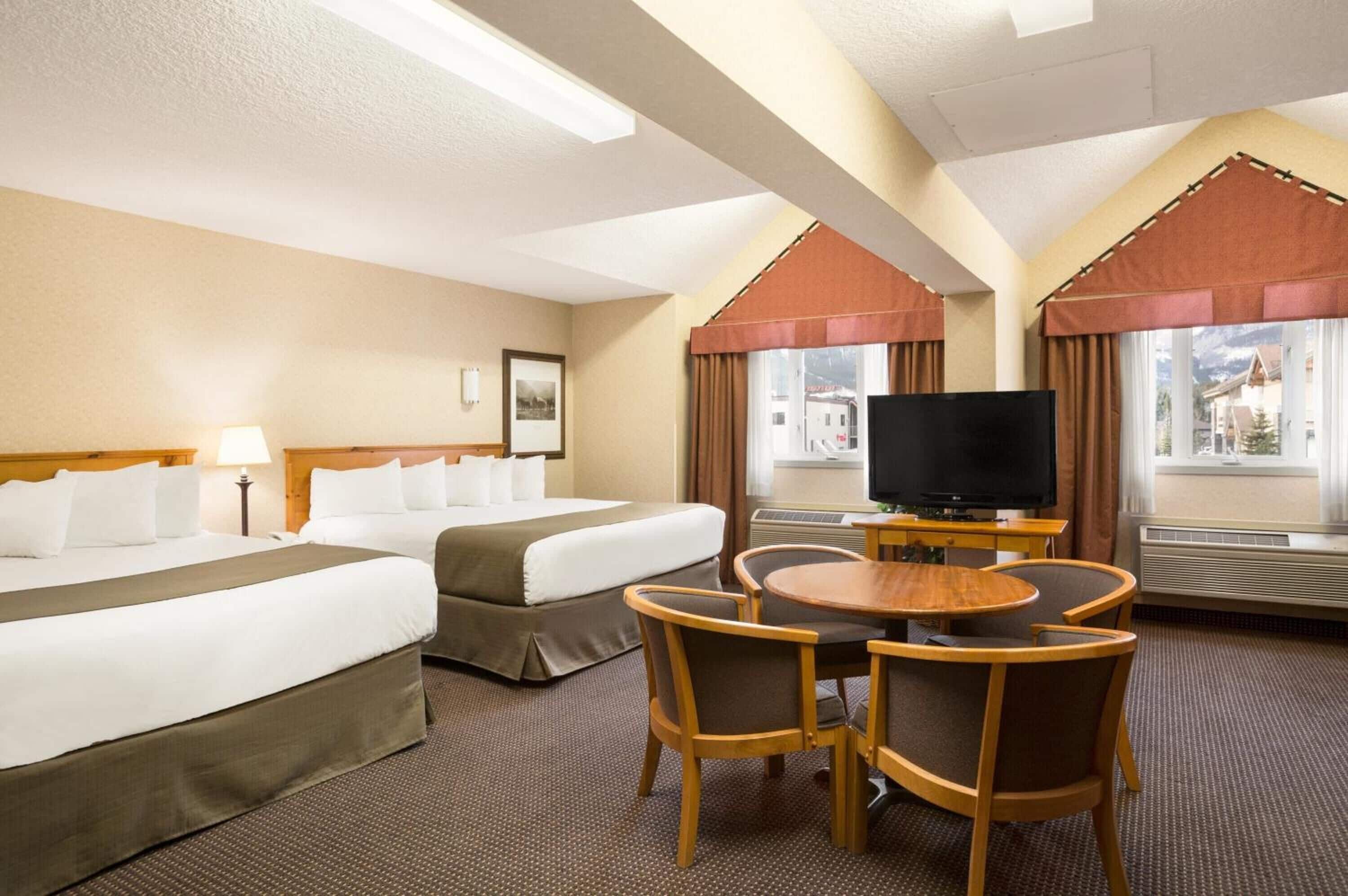 A hotel room with two beds, a TV, a dining table with chairs, and large windows with curtains.