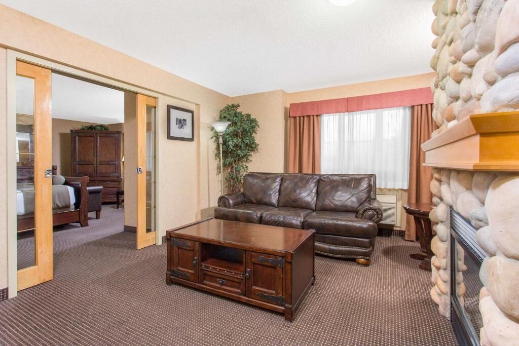 A hotel room with a leather sofa, a wooden coffee table, a stone fireplace, and a view of the adjacent room.