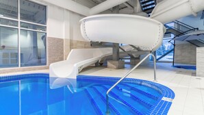 An indoor swimming pool with a slide, a staircase, and a view of a snowy landscape through large windows.