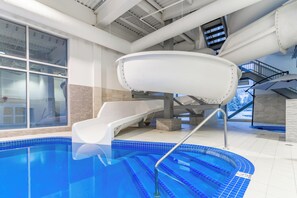 An indoor swimming pool with a slide, a staircase, and a view of a snowy landscape through large windows.