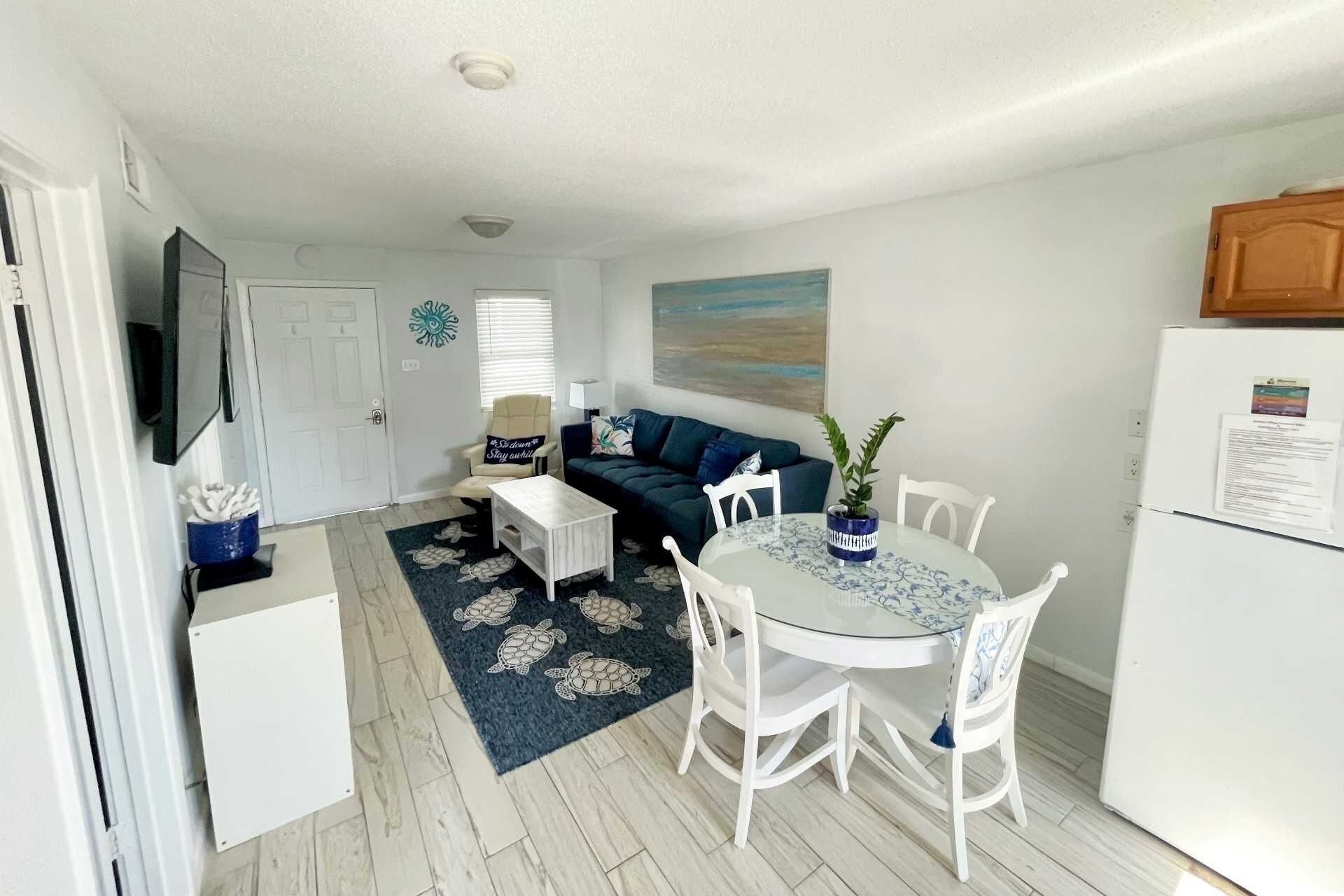 Un espace de vie compact comprenant un canapé, une table à manger avec des chaises, un téléviseur fixé au mur et un réfrigérateur.