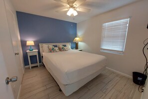 A bedroom with a bed, two bedside tables, a ceiling fan, a window with blinds, and a door.