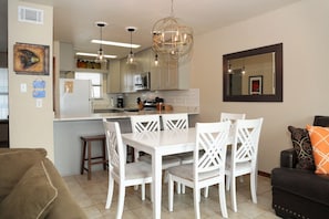 A modern kitchen with white cabinets, a central island, pendant lights, and a dining area with white chairs. - Ocean Reef 702 3 Bedroom Townhouse by RedAwning (Gulf Shores)