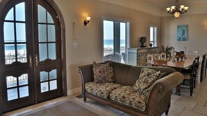 Ein geräumiges Wohnzimmer mit einer großen Holztür, einer Couch, einem Essbereich mit Stühlen und Blick auf den Ozean.