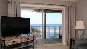A room with a TV on a wooden cabinet, a lamp, and a view of the beach through a sliding glass door.