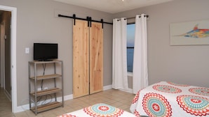 A bedroom with a bed, a TV on a stand, a ceiling fan, and a view of the sea through a window.