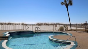 A swimming pool with a curved edge, surrounded by a sandy area and a wooden fence.