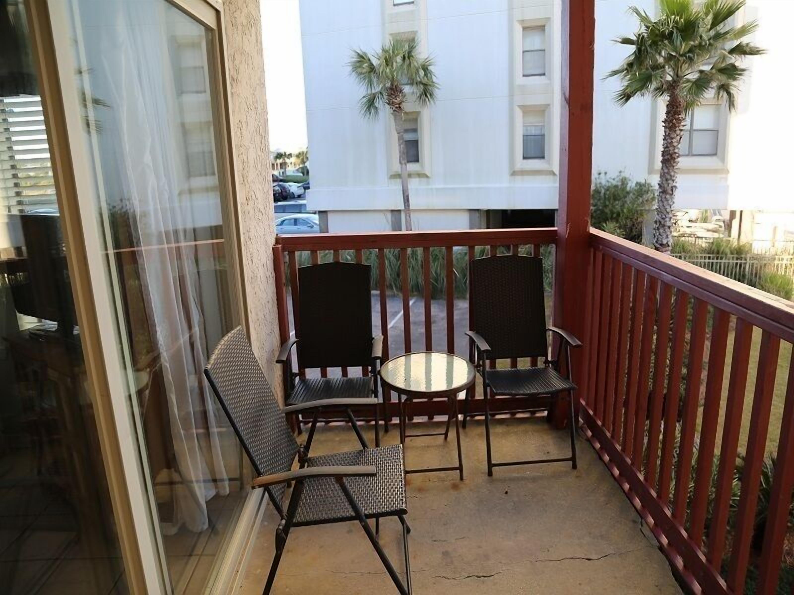 Un balcon avec deux chaises et une petite table, donnant sur une rue bordée de palmiers et sur un bâtiment blanc.