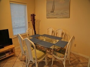 Un comedor con una mesa de vidrio y sillas blancas, un televisor sobre un mueble bajo, y un cuadro de un velero en la pared.