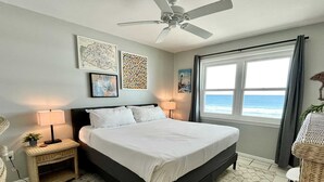 A bedroom with a bed, two bedside tables, a ceiling fan, a window with a view of the ocean, and artwork on the walls. - Harbor House B4 2 Bedroom Condo by RedAwning (Gulf Shores)