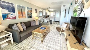 A living room with a sofa, coffee table, and TV. There are posters on the wall and a kitchen in the background. - Harbor House B4 2 Bedroom Condo by RedAwning (Gulf Shores)