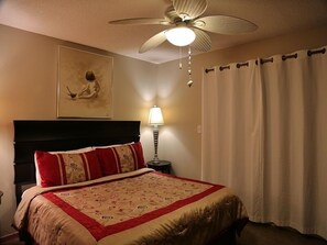 A hotel room with a bed, a ceiling fan, a nightstand with a lamp, a painting on the wall, and a curtain. - H Oz-Toto 3 Bedroom Home by RedAwning (Gulf Shores)