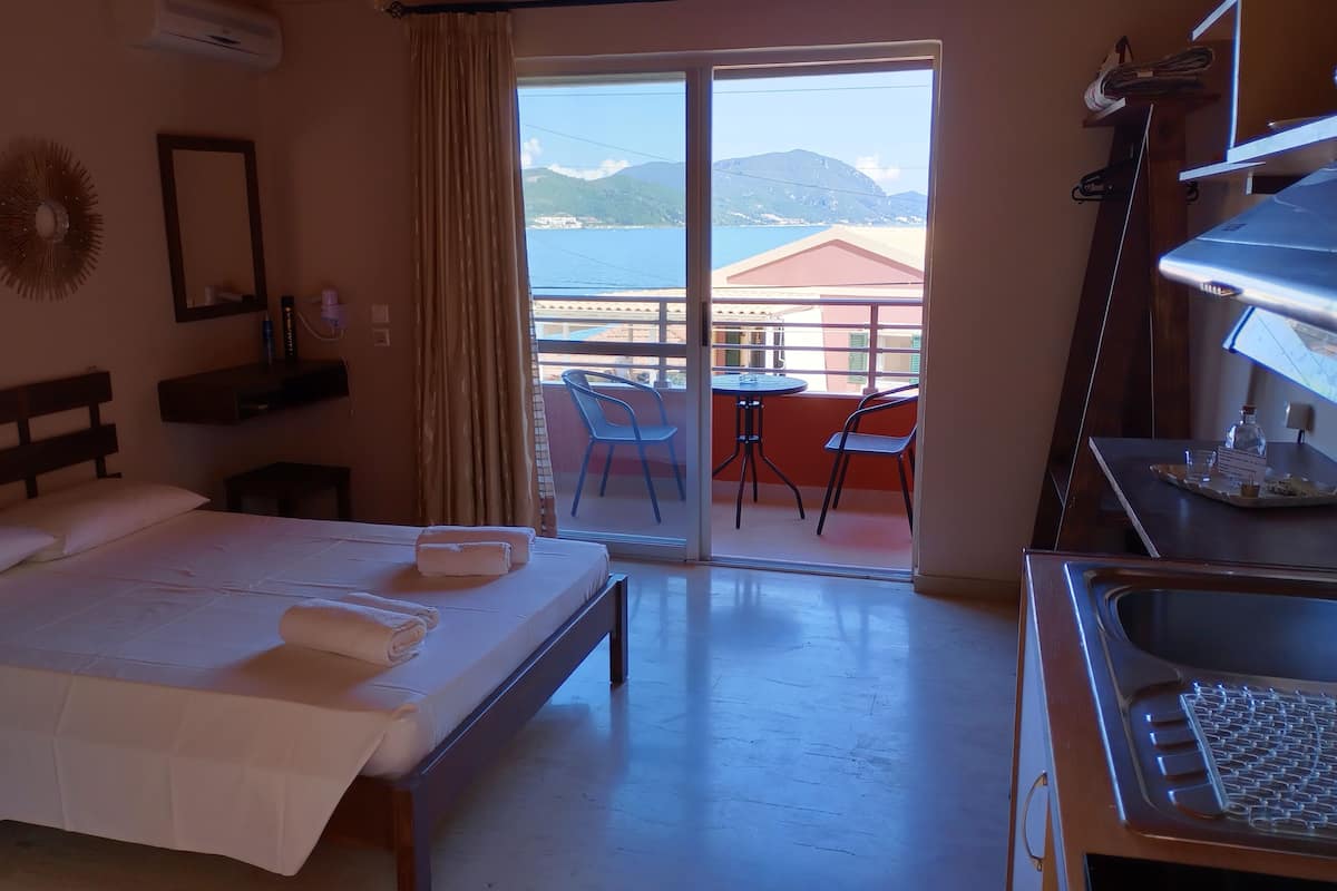 A hotel room with a bed, a desk, a chair, and a balcony with a view of the sea and mountains.