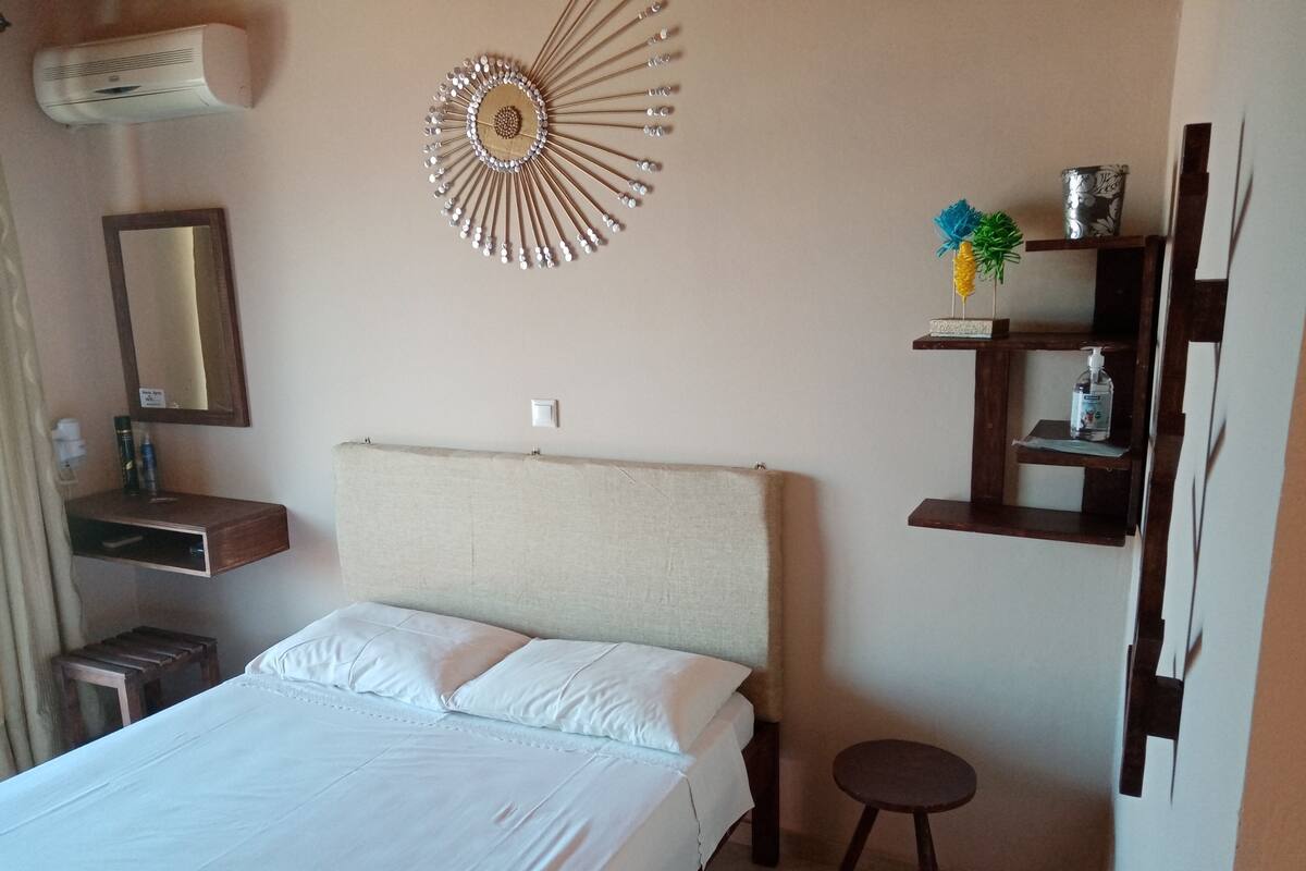A hotel room with a bed, a bedside table, a mirror, a wall-mounted decorative item, and a small shelf with toiletries.