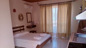 Habitación de hotel con cama, mesita de noche, espejo, equipo de aire acondicionado, ventana con cortinas y una zona de kitchenette.