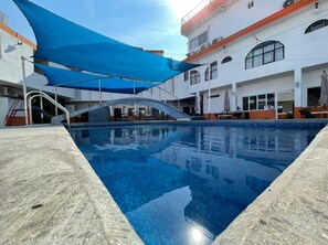 Une piscine surmontée d’une voile d’ombrage bleue, entourée d’un bâtiment doté de nombreux balcons et fenêtres.