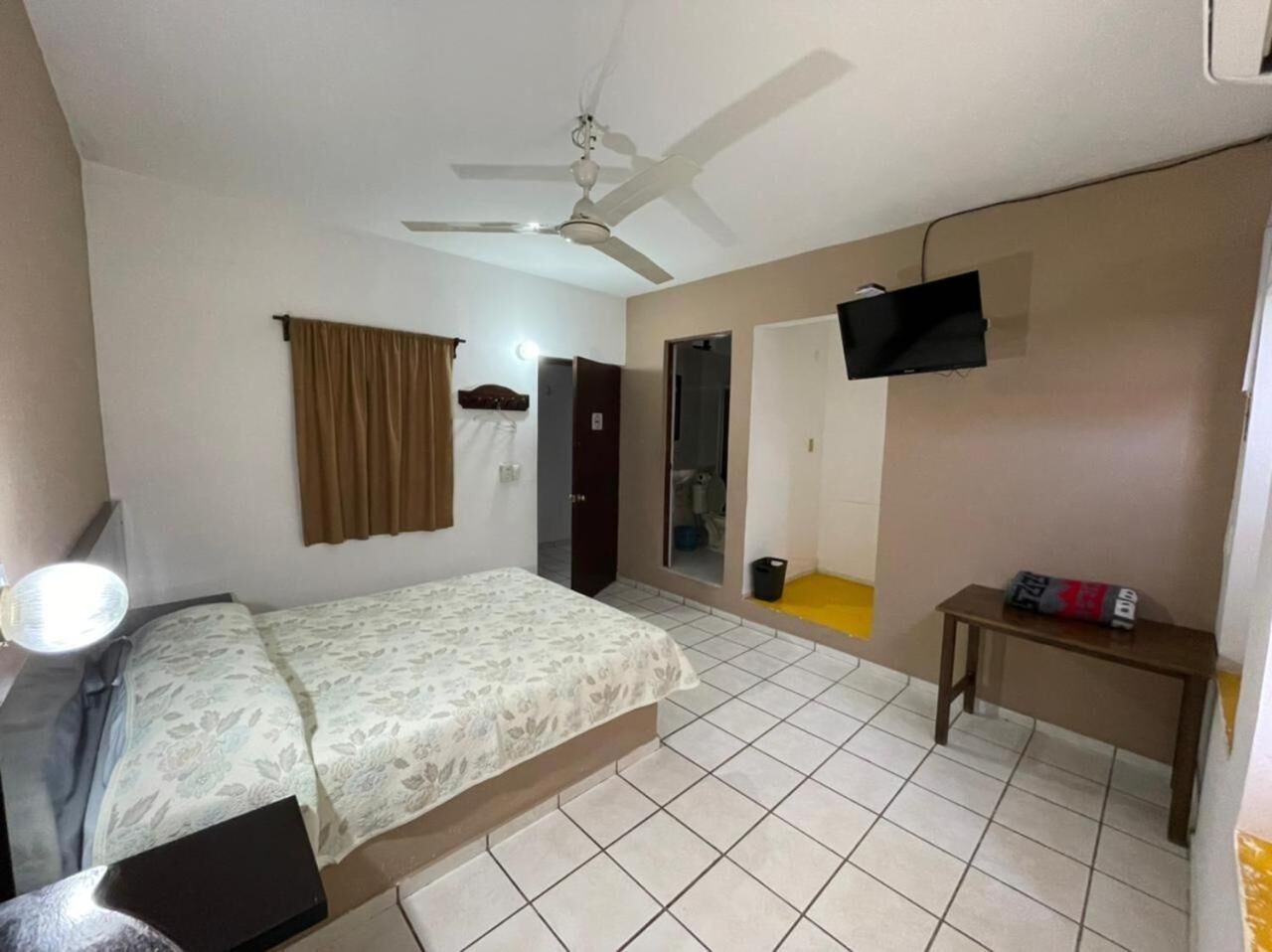 A bedroom with a bed, a ceiling fan, a TV mounted on the wall, a small table, and a door leading to another room.