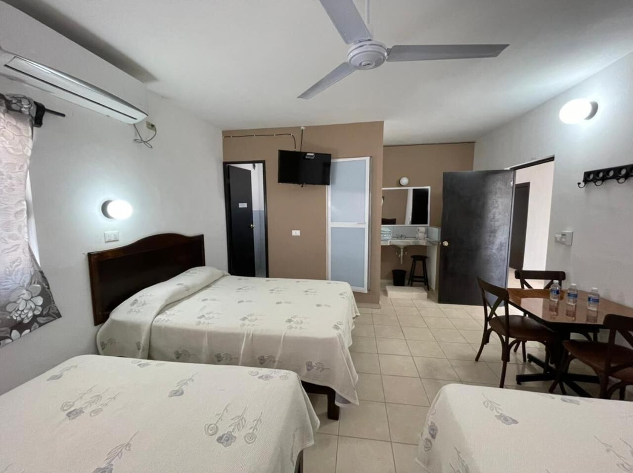 A hotel room with two beds, a dining table with chairs, a ceiling fan, and a TV mounted on the wall.