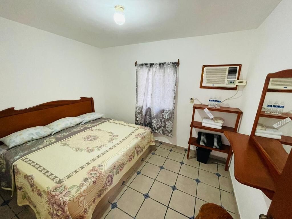 A bedroom with a bed, a window with curtains, an air conditioning unit, and a small desk with a chair.