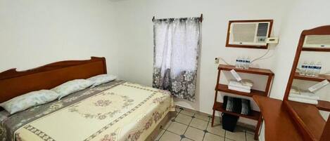 A bedroom with a bed, a window with curtains, an air conditioning unit, and a small desk with a chair.