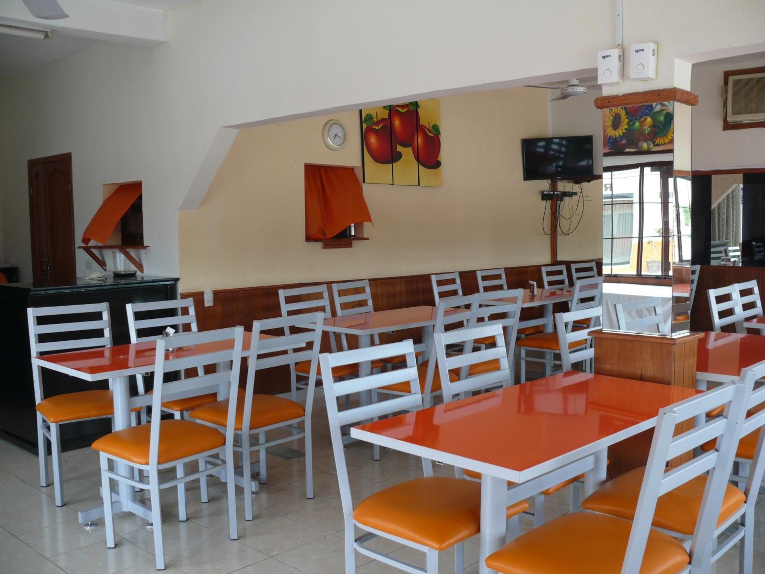 Um restaurante com mesas brancas e cadeiras laranja, um relógio e pinturas de frutas nas paredes.