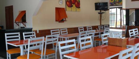 Um restaurante com mesas brancas e cadeiras laranja, um relógio e pinturas de frutas nas paredes.
