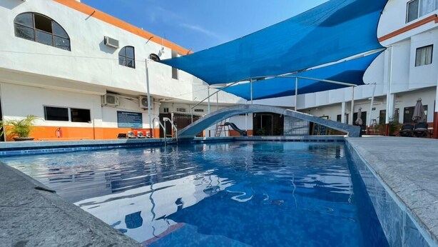 A swimming pool with a blue shade sail overhead, surrounded by buildings with orange and white facades. - Hotel Moreno (Ciudad Valles)