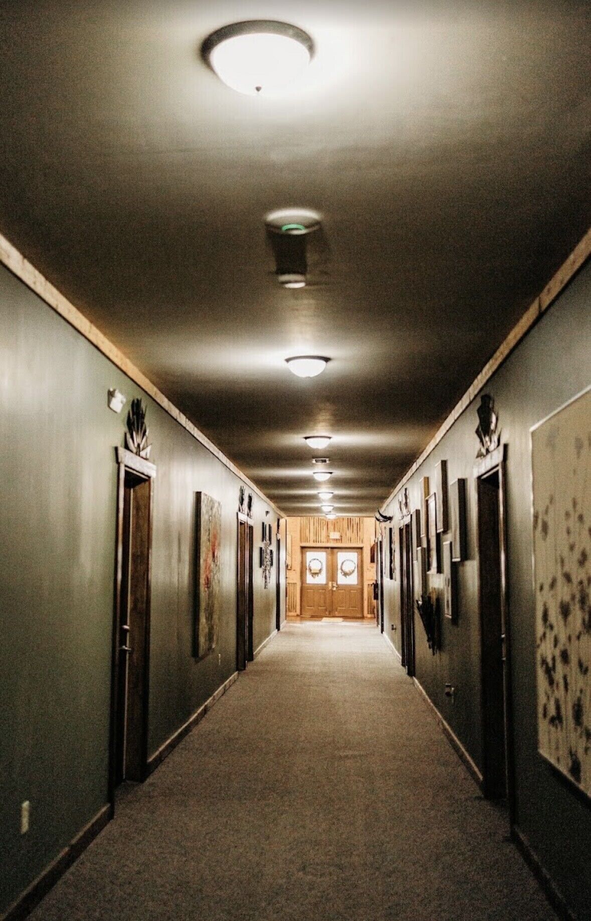 Un long couloir étroit, doté d’un sol recouvert de moquette, de nombreuses portes de chaque côté et d’une porte en bois au bout.
