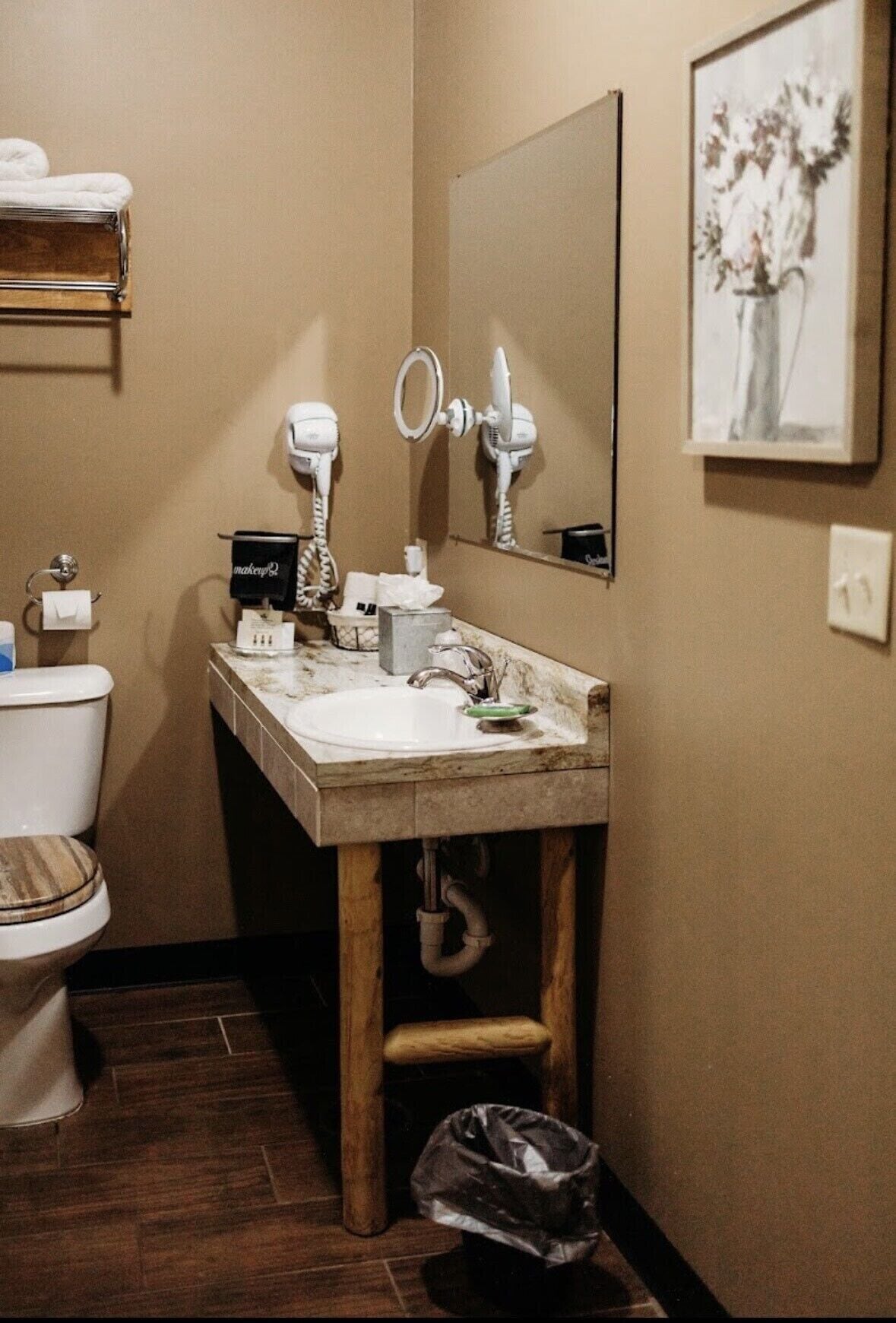 A bathroom with a wooden vanity, a white sink, a hairdryer, a mirror, and a framed artwork on the wall.