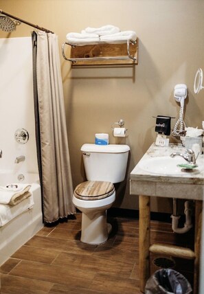 A bathroom with a wooden toilet, a shower curtain, a towel rack with towels, a toilet paper holder, a hairdryer, and a sink with a mirror. - Big Horse Inn and Suites (Lewistown)