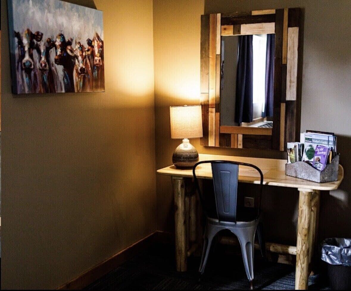 A hotel room with a wooden desk, a chair, a lamp, a mirror, and a painting on the wall.