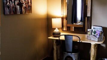 A hotel room with a wooden desk, a chair, a lamp, a mirror, and a painting on the wall.