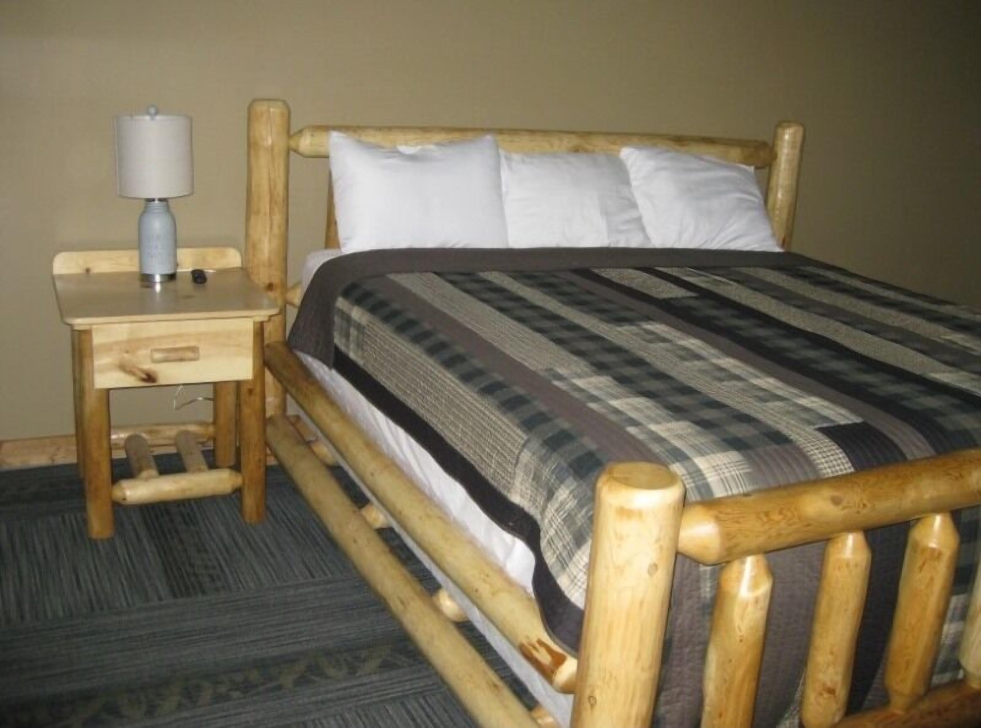 Una cama de madera con cubrecama a cuadros, una mesita de noche de madera con una lámpara, y suelo alfombrado.