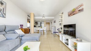 A modern living room with a grey sofa, a white coffee table, a TV, and a kitchen in the background.