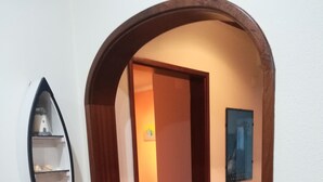 A hallway with a wooden archway leading to a bedroom. There is a shelf with a small item on the left and a framed picture on the wall to the right. - Sesimbra Holiday Apartment (Sesimbra)