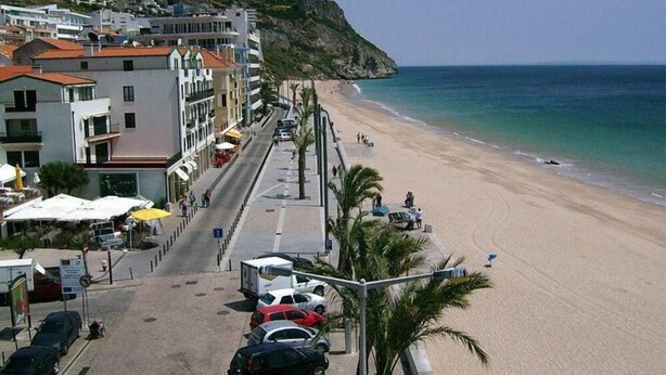 A coastal street with buildings, parked cars, and a beach. - Sesimbra Holiday Apartment (Sesimbra)